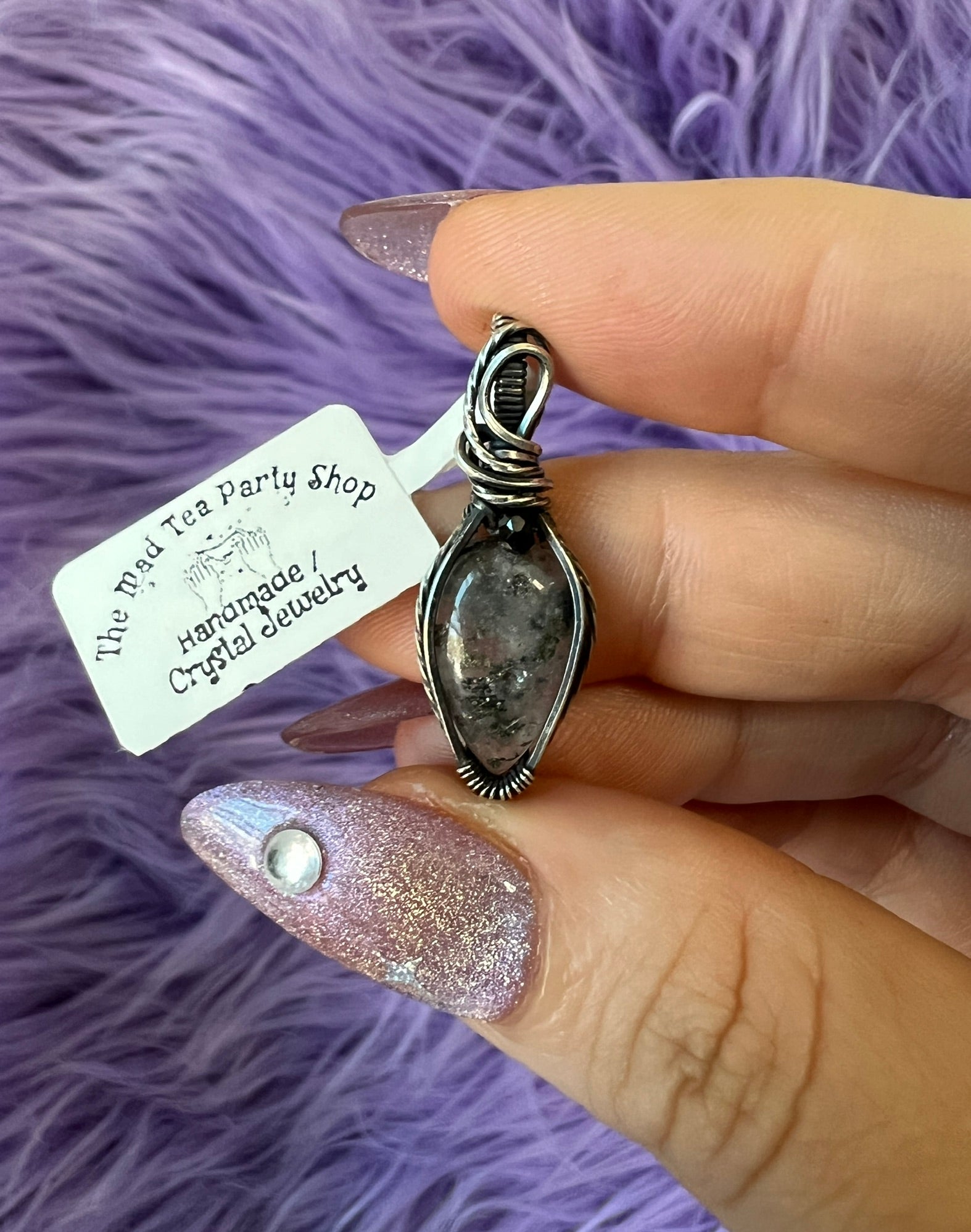 Hand holding a silver pendant with a tag from The Tea Party Shop on a purple fluffy background